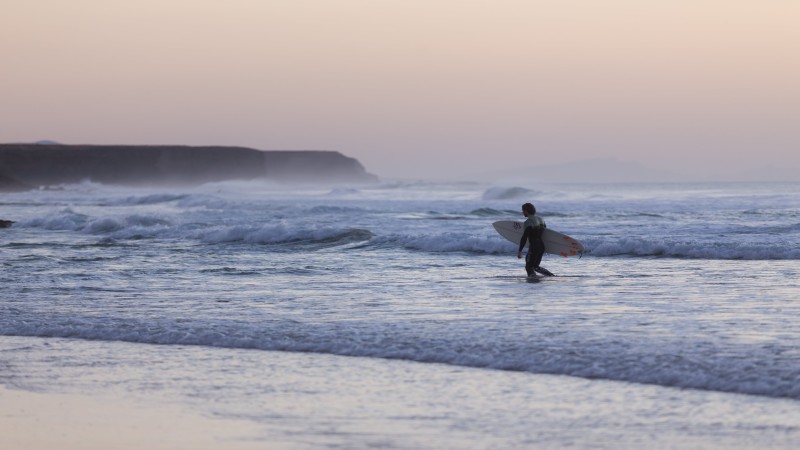 Surfeur entrant dans l'eau à El Cotillo au coucher du soleil, avec de douces vagues et des tons violets dans le ciel, reflétant la tranquillité du moment.